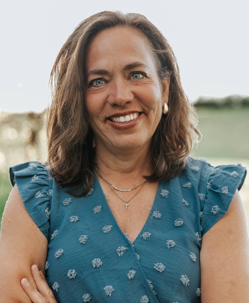 Smiling woman with shoulder-length brown hair wearing a blue patterned blouse outdoors.